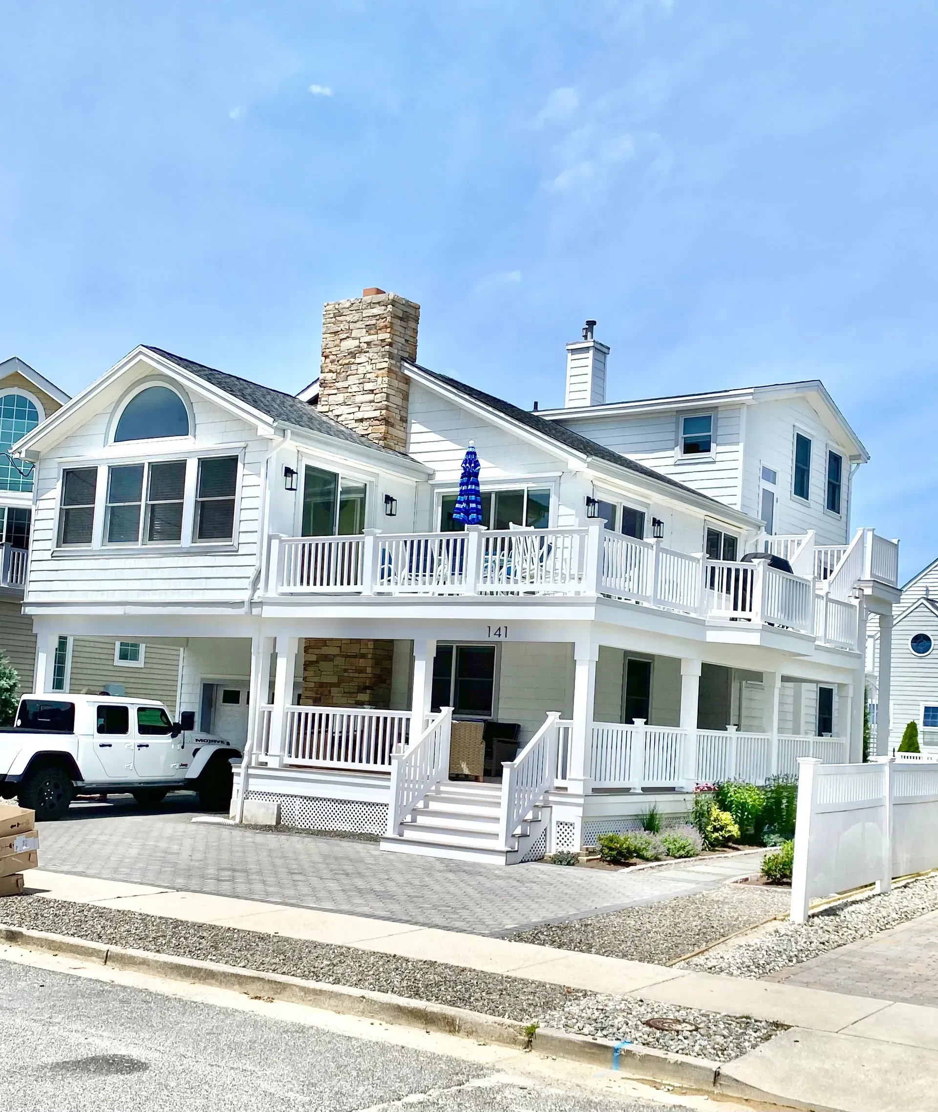 A white, two-story coastal home with a stone chimney, wrap-around porch, and a white pickup truck parked in the driveway.