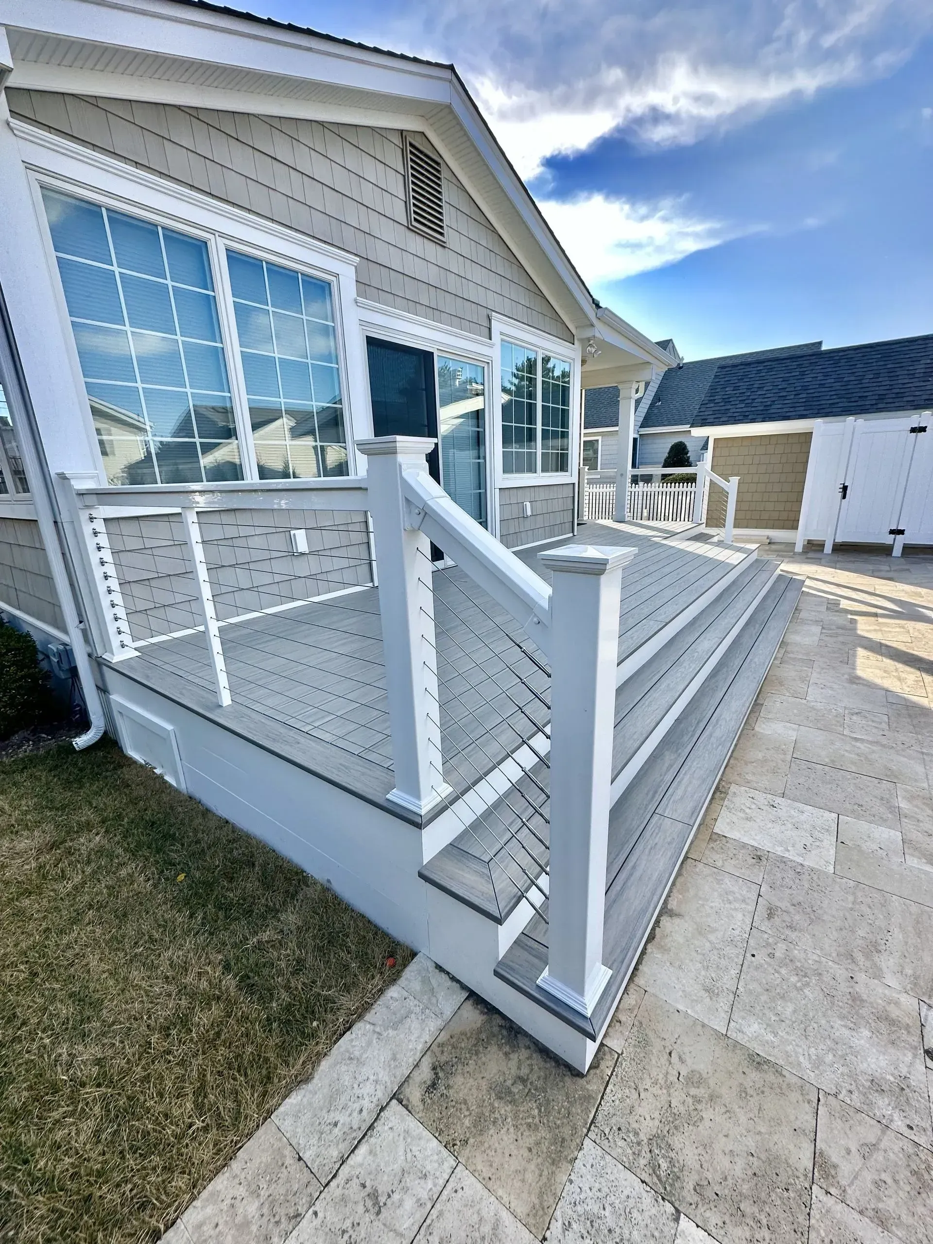 A gray wooden deck with white railing and stairs leads to a house with large windows under a bright blue sky.