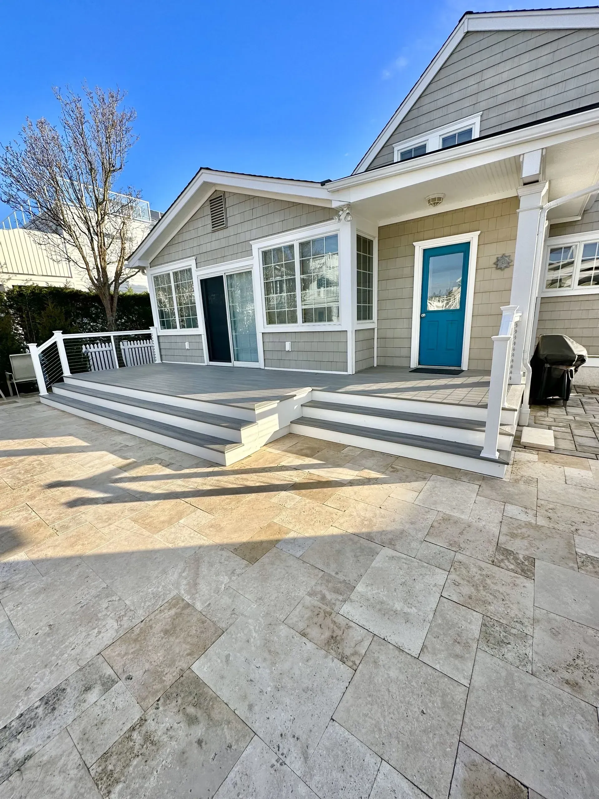 Exterior view of a light-gray house with a bright blue door, steps leading to a deck, and a stone patio.