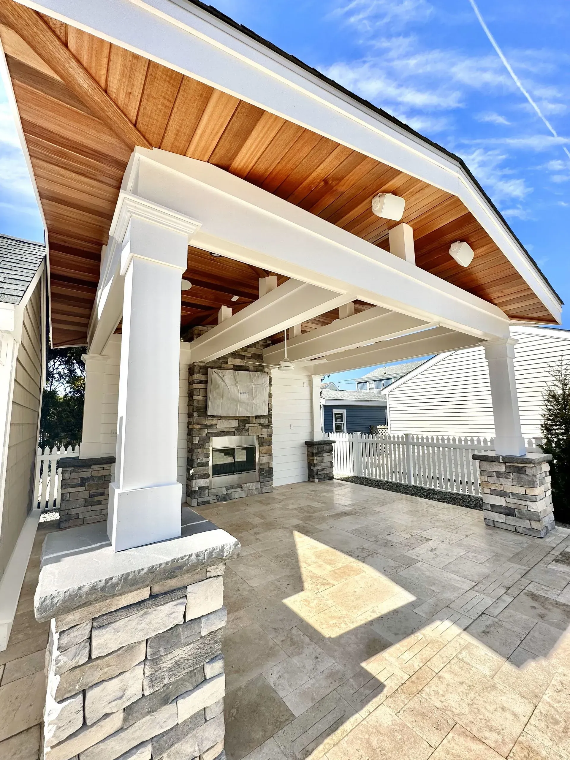 An outdoor covered patio with a stone fireplace, white columns, and a natural wood ceiling under a bright blue sky.
