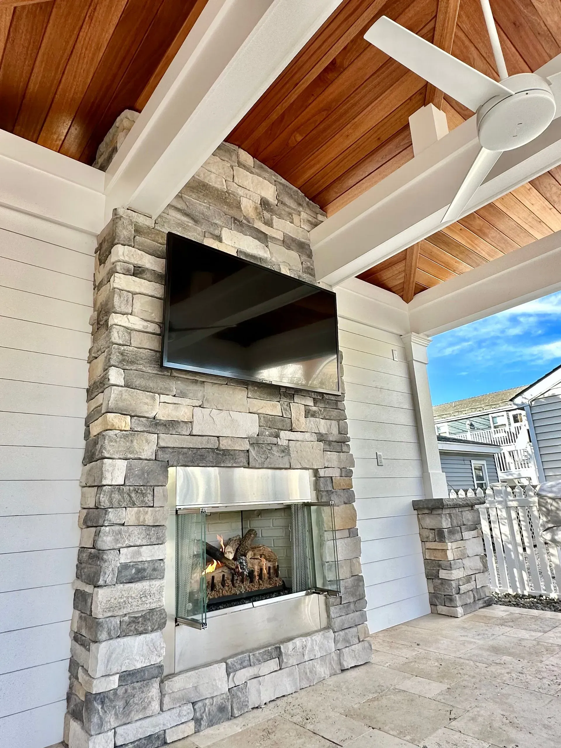 A stone outdoor fireplace and mounted TV on a covered patio with white shiplap walls and a wood-paneled ceiling.
