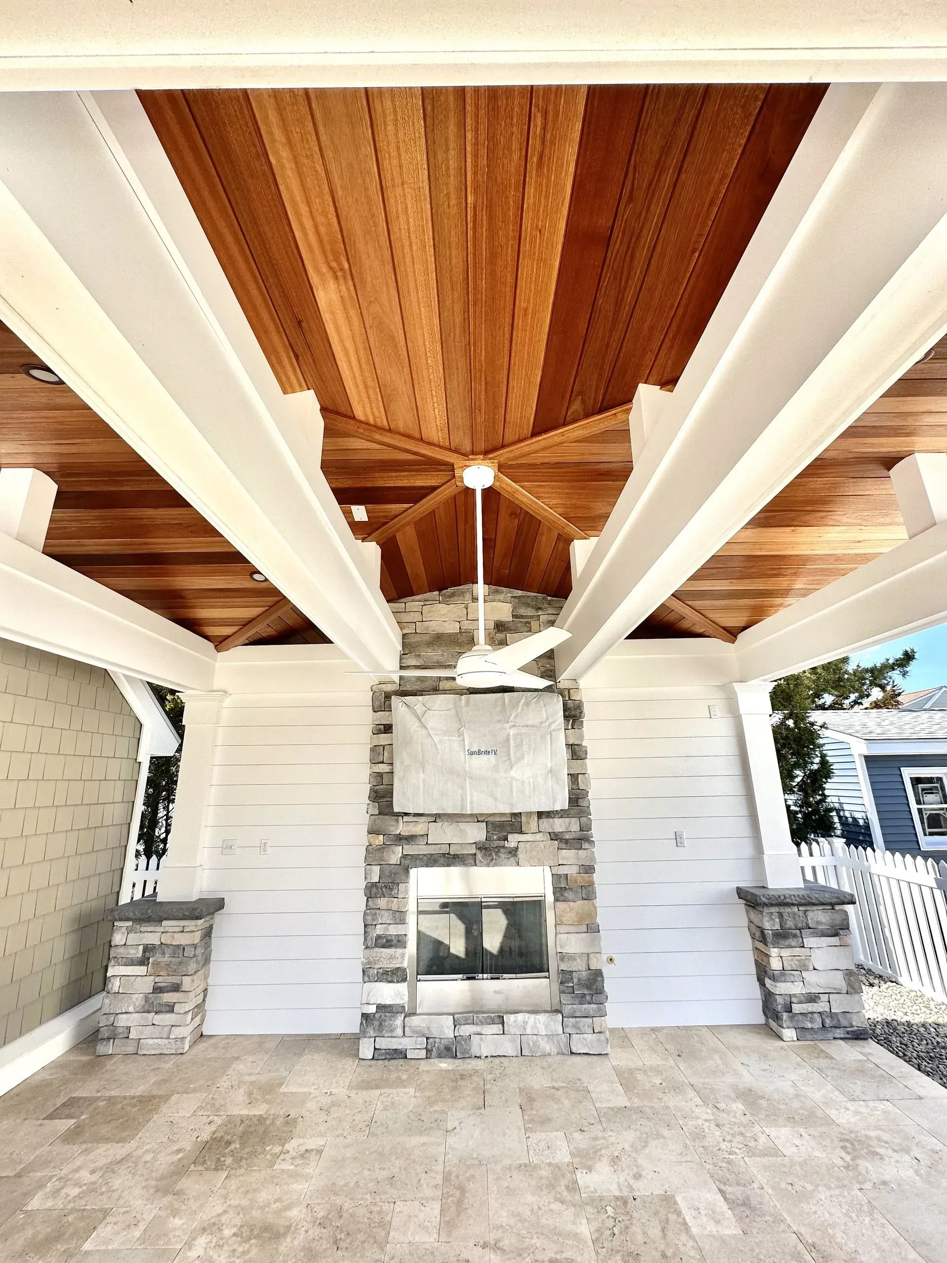An outdoor covered patio with a stone fireplace, light tile flooring, white support pillars, and a wood-paneled ceiling.