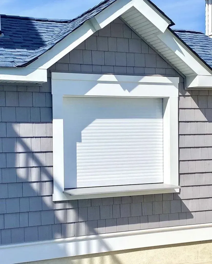 A window with white closed security shutters set into a gray shingled exterior wall under a gabled roof.