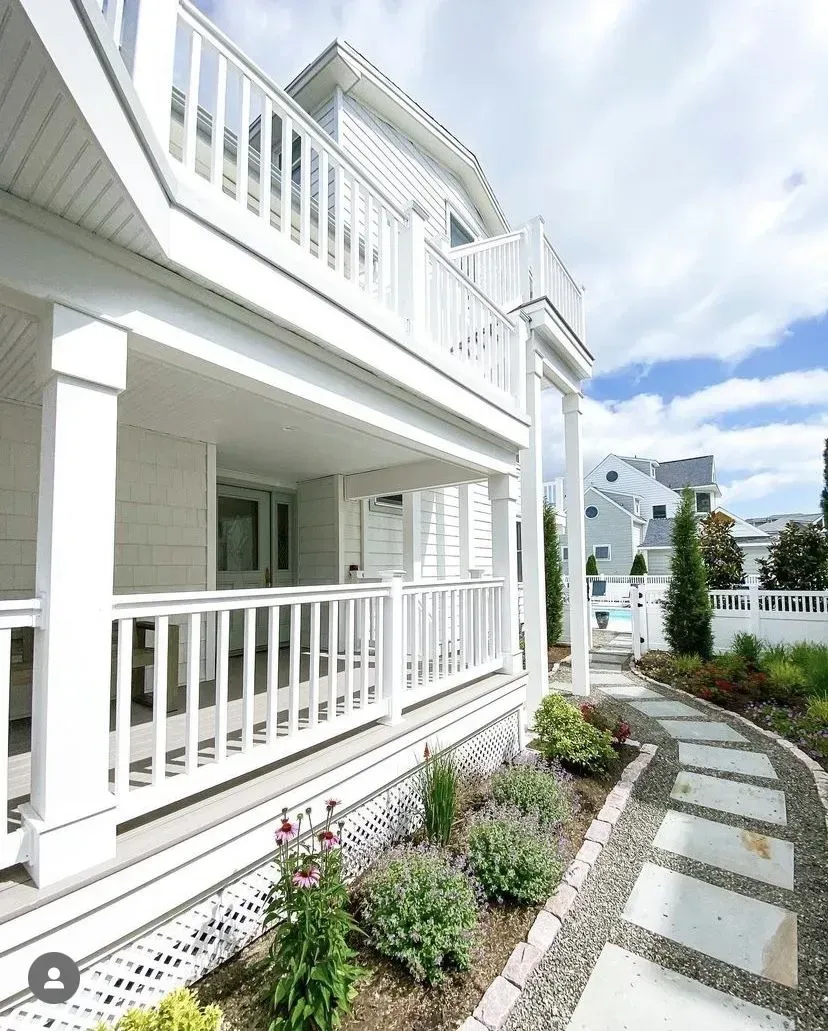 White two-story house with a wraparound porch and balcony, featuring a stone path leading through a garden.