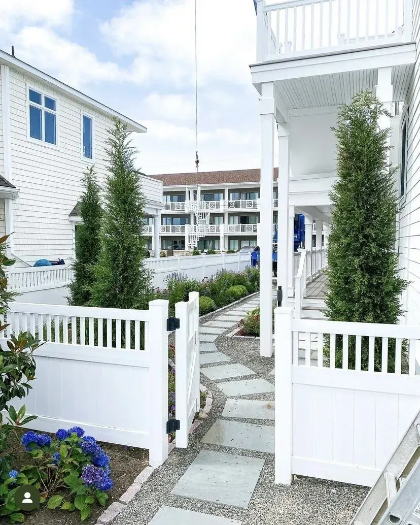 A white picket gate leads to a stone walkway between two white buildings with tall evergreen trees and blue hydrangeas.