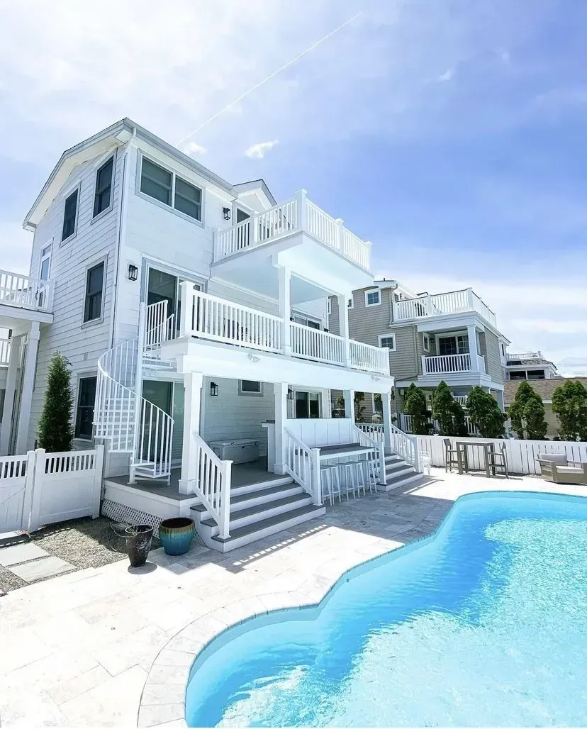 A light blue multi-story beach house with balconies, stairs, and an outdoor bar overlooking a curved swimming pool.
