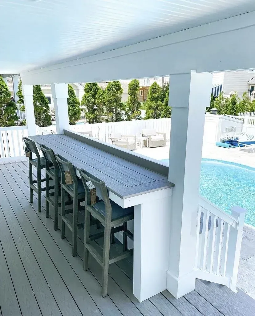 A gray bar counter with four wooden stools on a covered outdoor deck overlooking a swimming pool.
