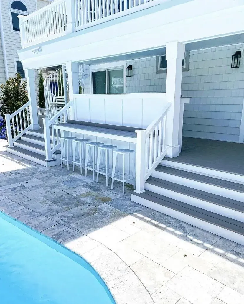 White outdoor patio bar with four tall stools, nestled under a deck next to a blue swimming pool.