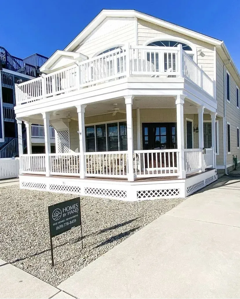 A two-story, light yellow beach house with white railings, a wrap-around porch, and a gravel yard under a clear blue sky.