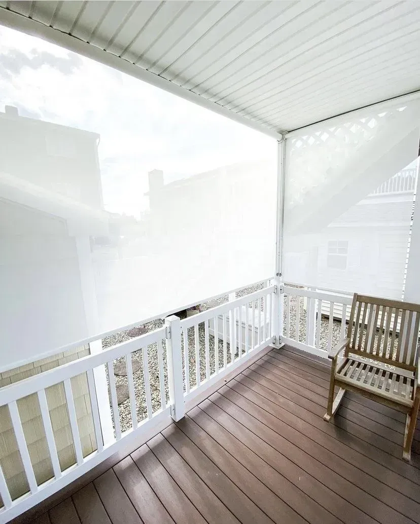 A covered outdoor balcony with wooden flooring, white railings, and translucent white privacy shades.