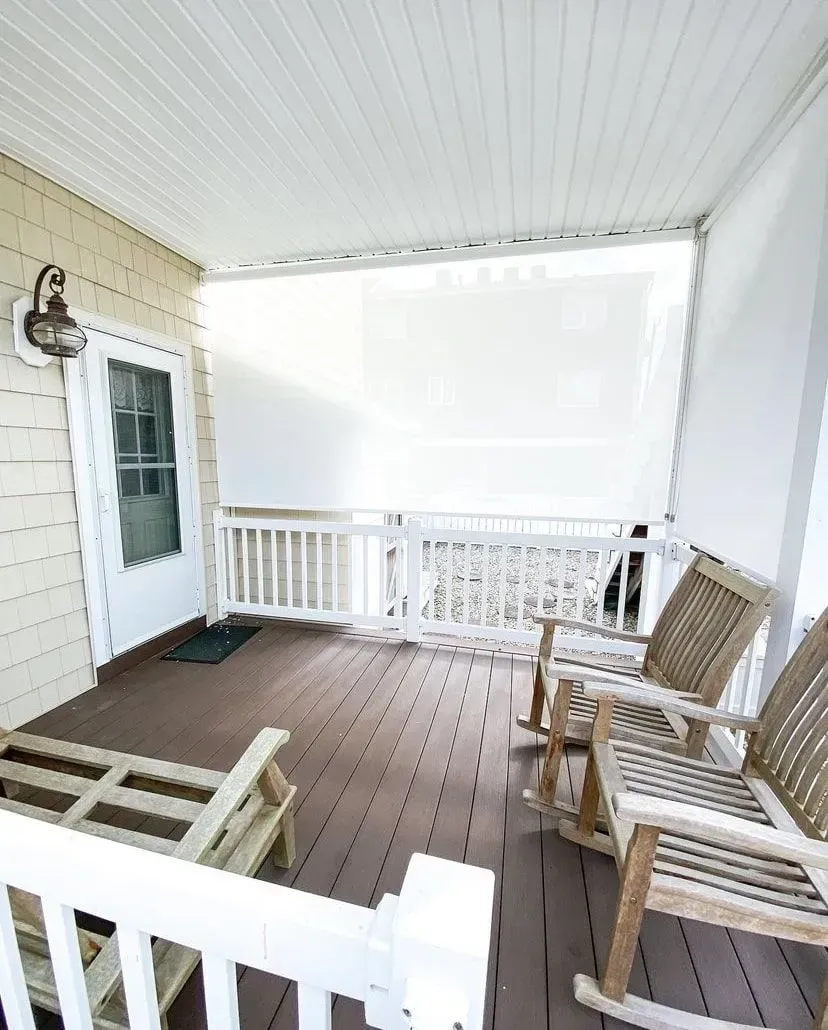A wooden deck featuring two rocking chairs, a bench, and a door leading inside, all sheltered by a white roof.
