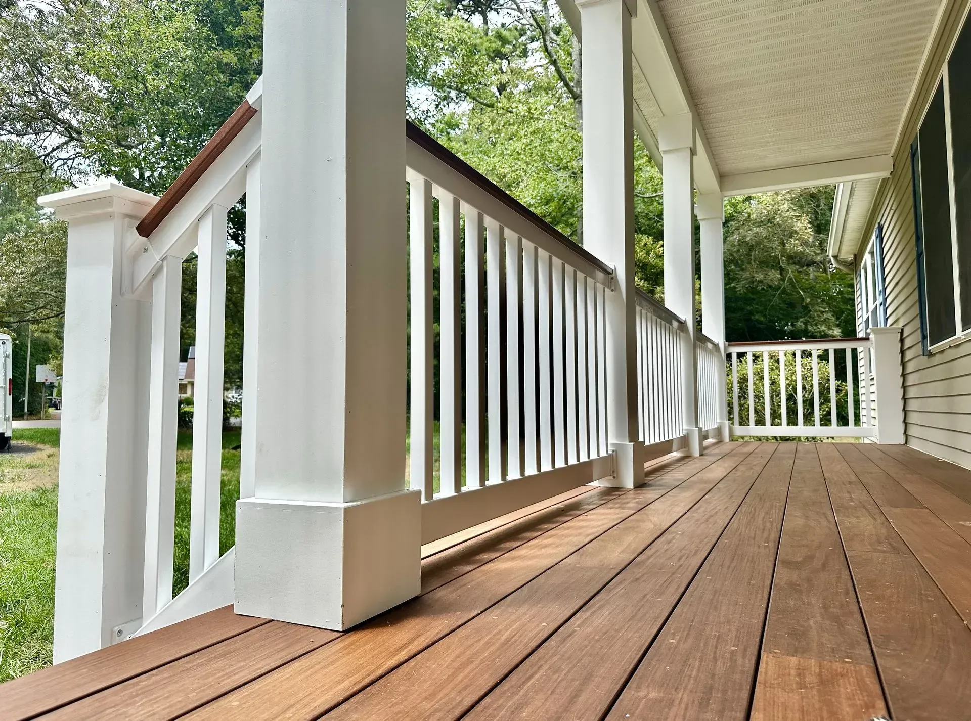 A wooden porch deck with a white railing and square support columns, viewed from a low angle along the length of the porch.