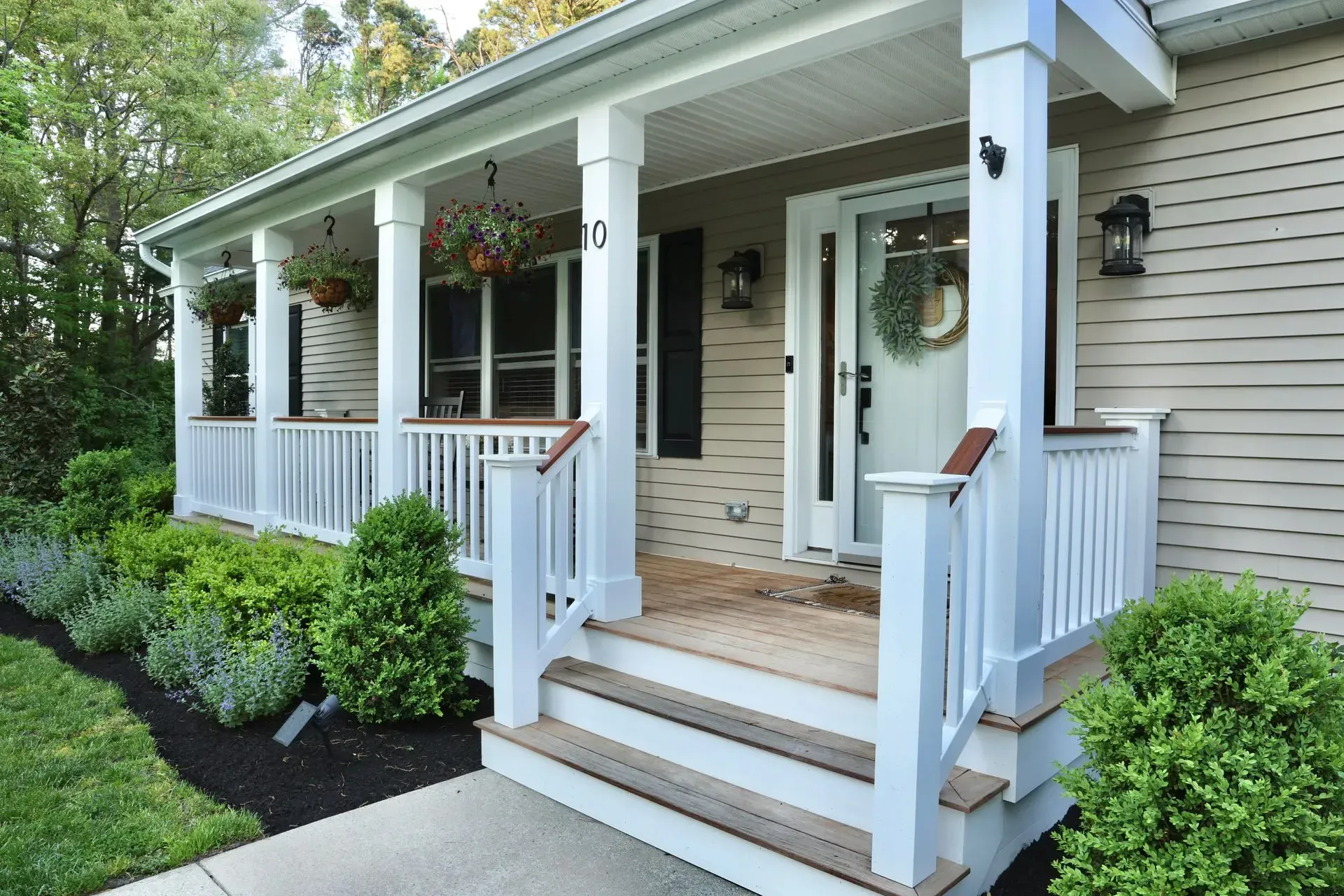 The front porch of a house with white railings, columns, a front door with a wreath, and landscaping in a mulch bed.
