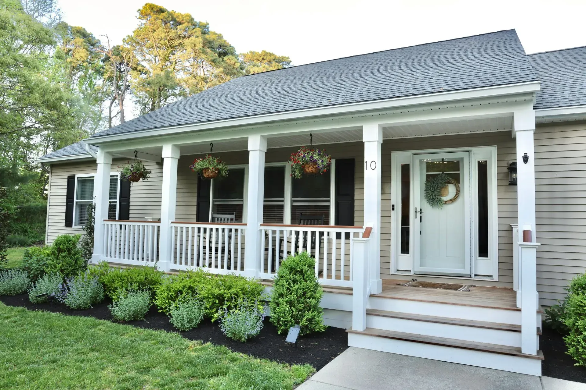 A light-tan, single-story house with a white front porch, black shutters, and well-manicured landscaping.