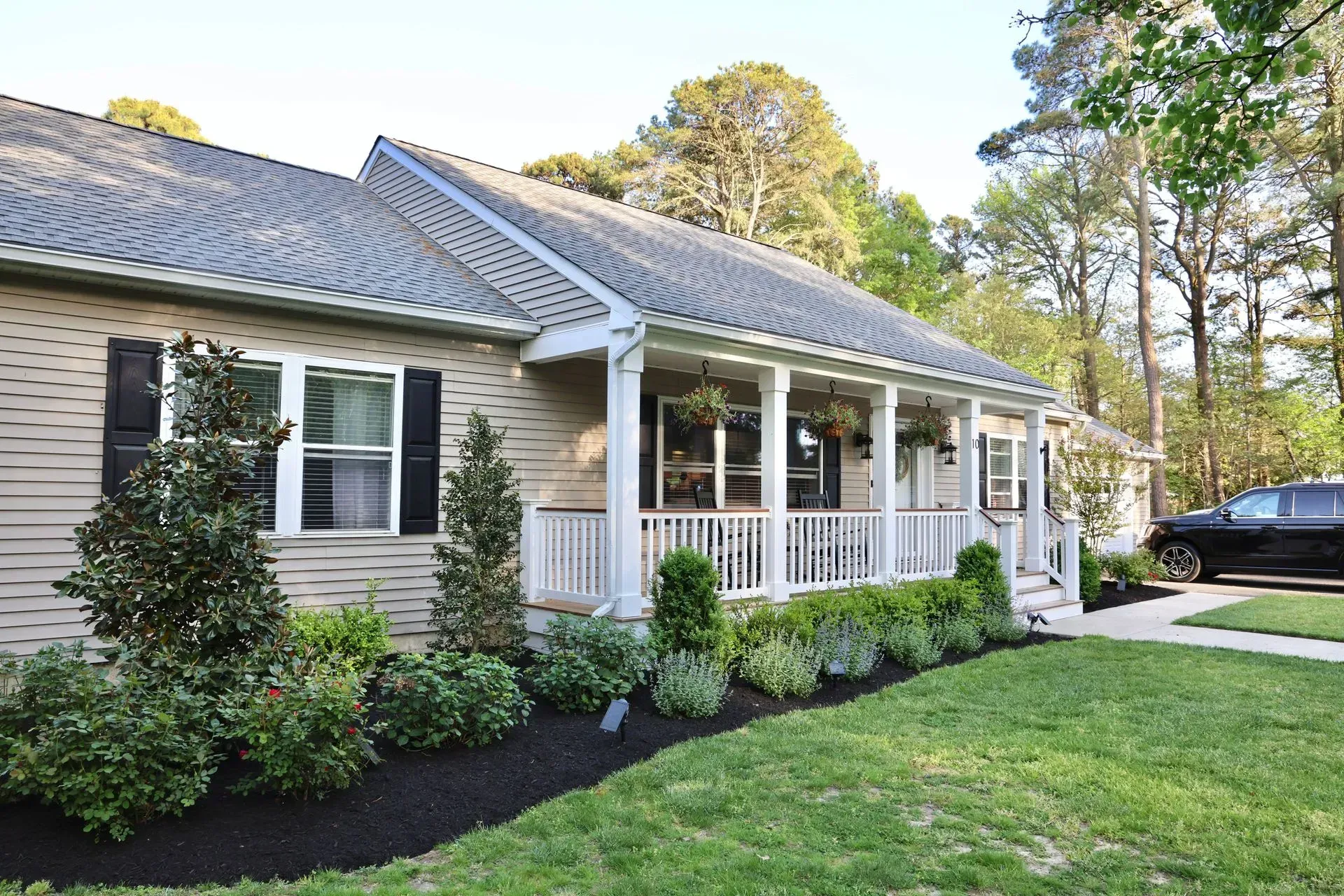 A beige, one-story suburban home with a front porch, white railings, and manicured landscaping under a bright sky.