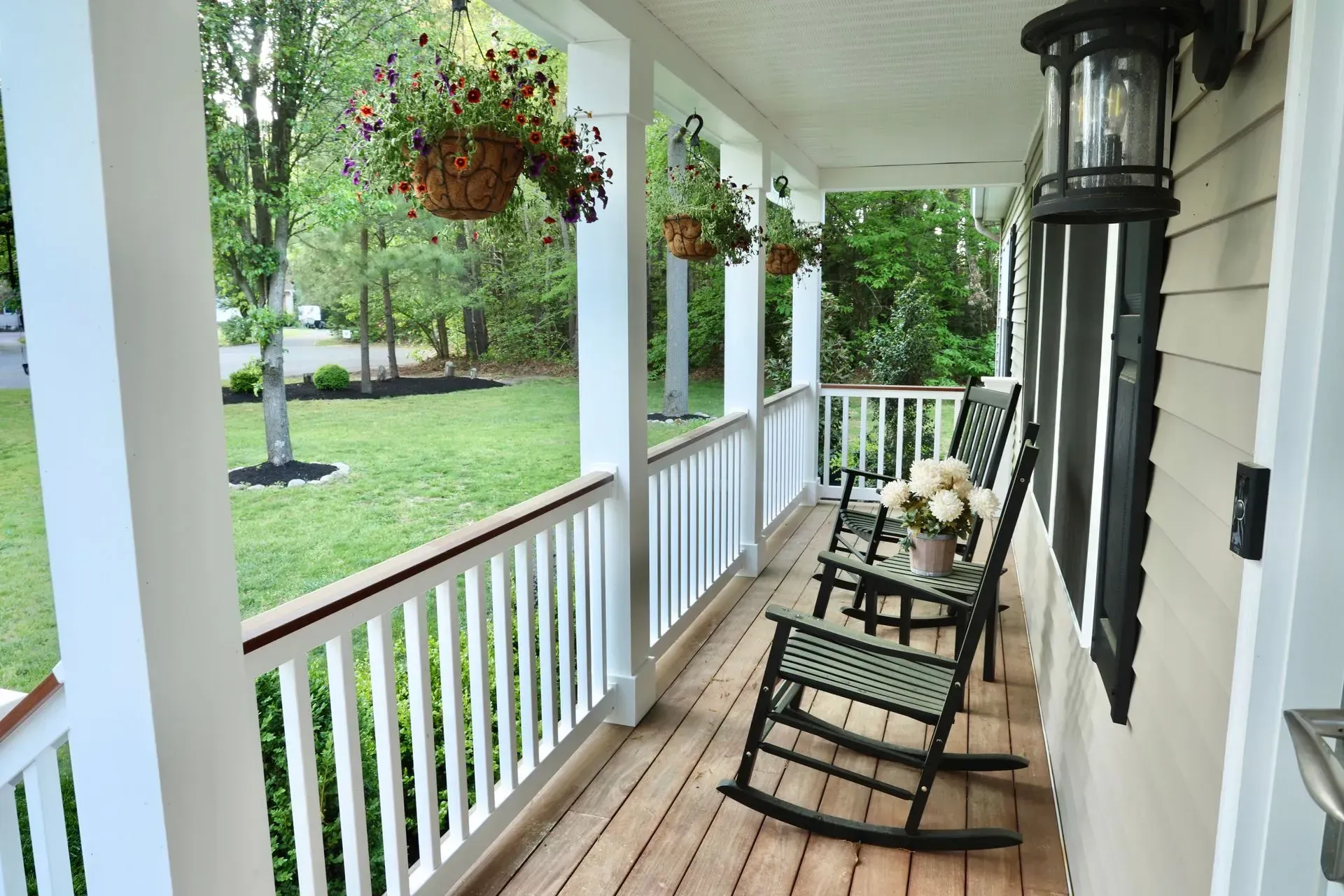 A front porch with wooden floors, white railings, two black rocking chairs, and hanging flower baskets.