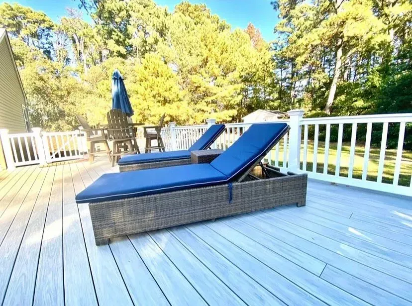 Two blue lounge chairs sit on a light-colored wooden deck in front of a white railing and trees under a sunny blue sky.