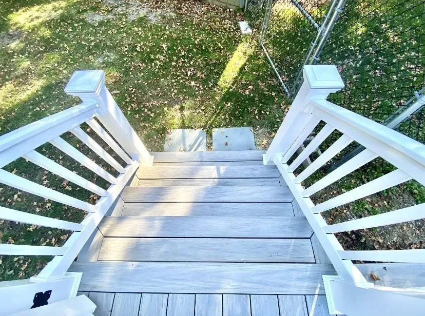 A set of wooden stairs with white railings leading down from a deck to a grassy yard covered in fallen leaves.