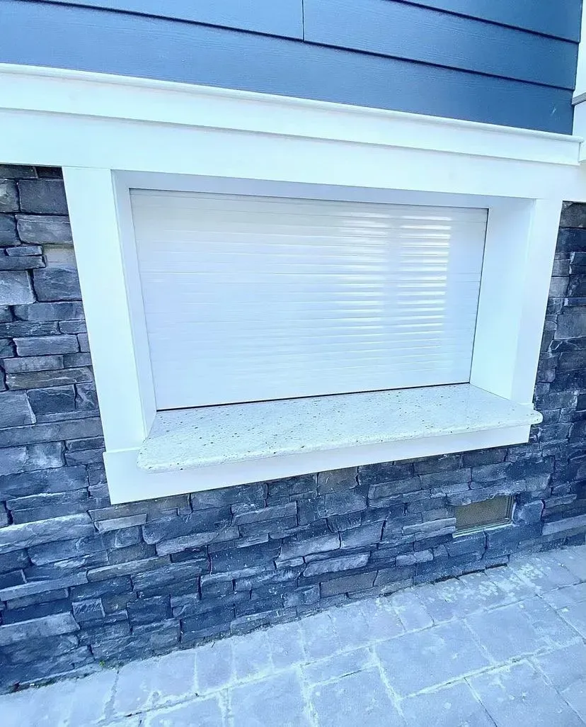 A recessed outdoor window with white trim, a closed shutter, and a granite sill set against a dark gray stone wall.