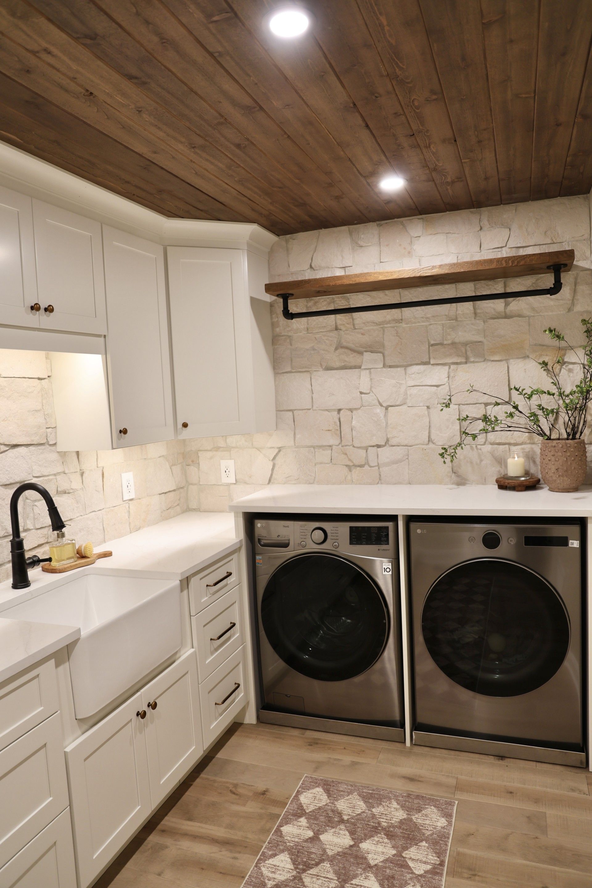 Modern laundry room with white cabinets, stone backsplash, black sink, and stacked washer-dryer under wood ceiling