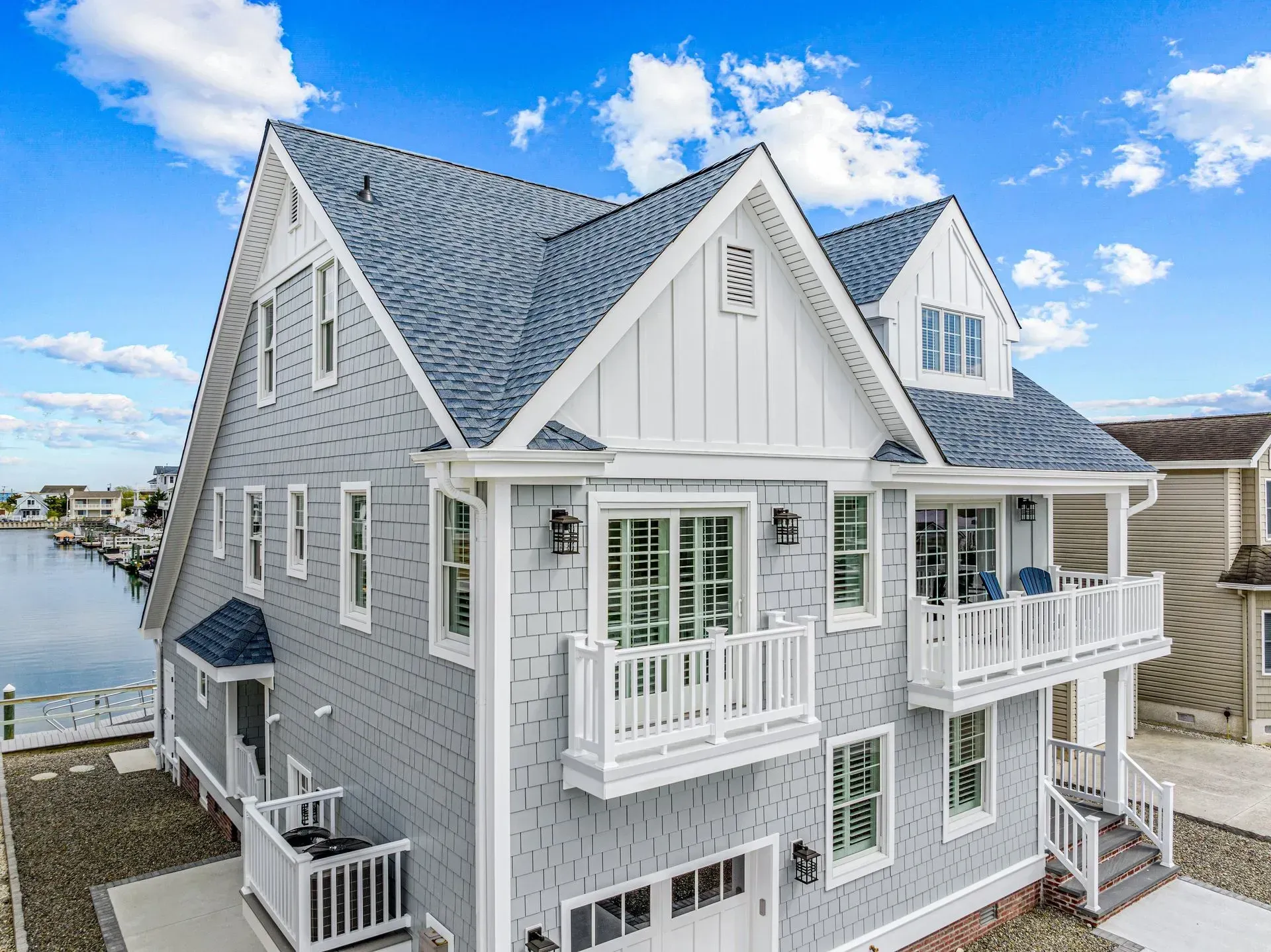 A three-story, grey shingled coastal house with a white trim, multiple balconies, and a blue roof near a canal.