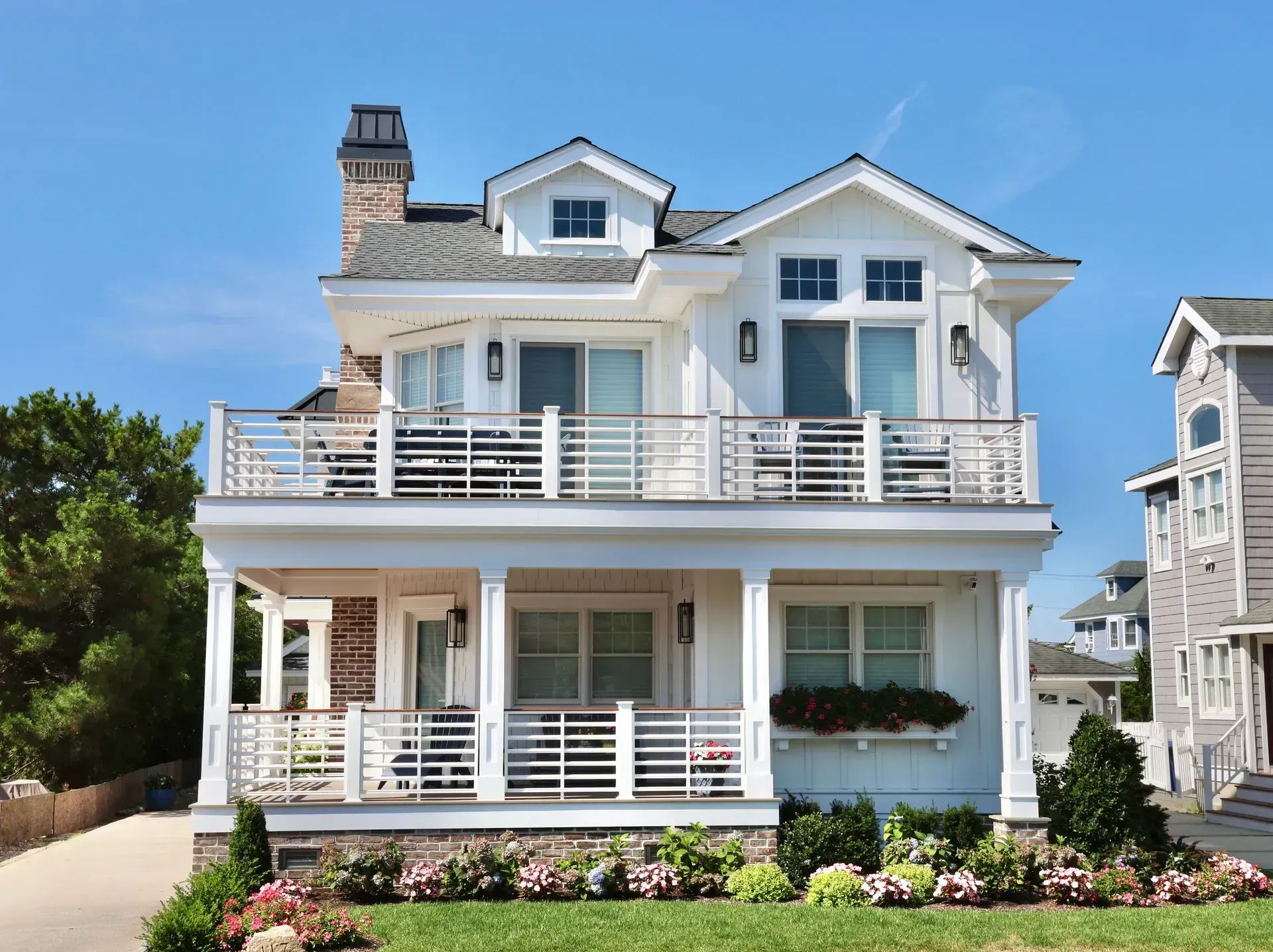 A two-story, white coastal-style house with a front porch, balcony, and a brick chimney under a clear blue sky.