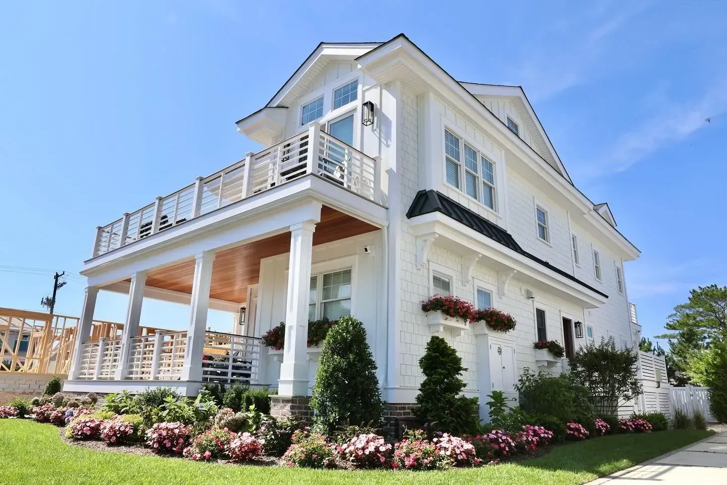 Two-story, white coastal-style house with a front porch, second-floor balcony, and manicured landscaping under a blue sky.