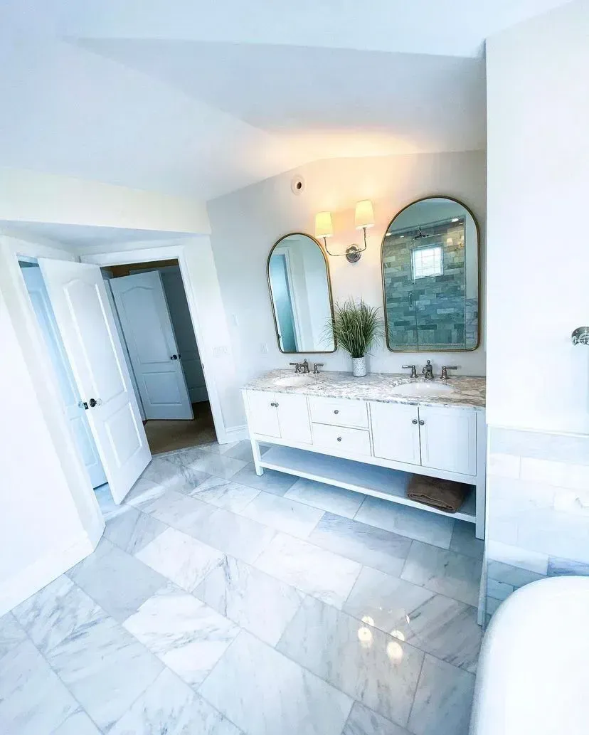 A bright bathroom featuring a white double vanity, two arched mirrors, marble tile flooring, and an open doorway.