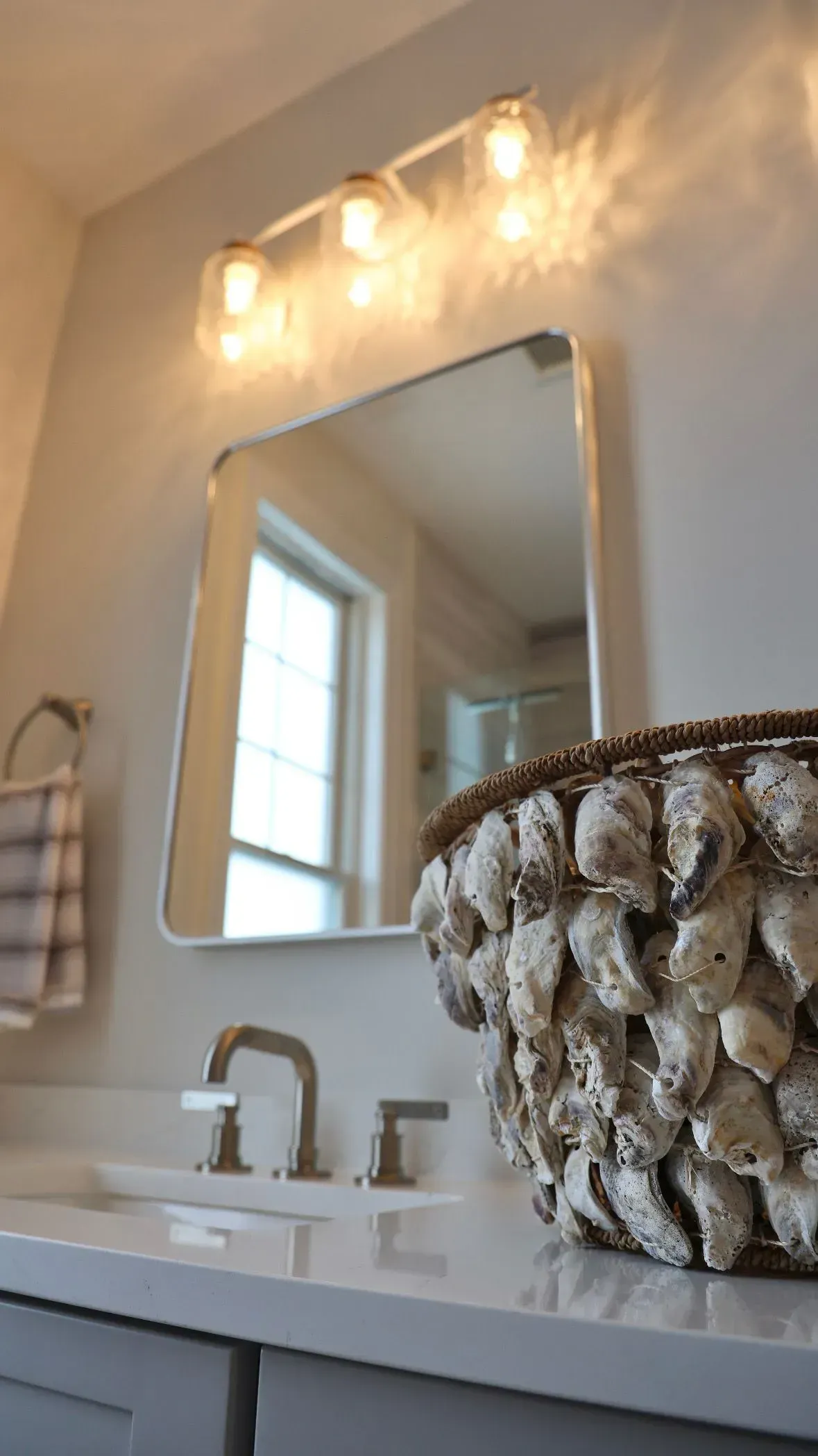 A bathroom vanity with a square mirror, a modern three-light fixture, and a decorative basket covered in oyster shells.