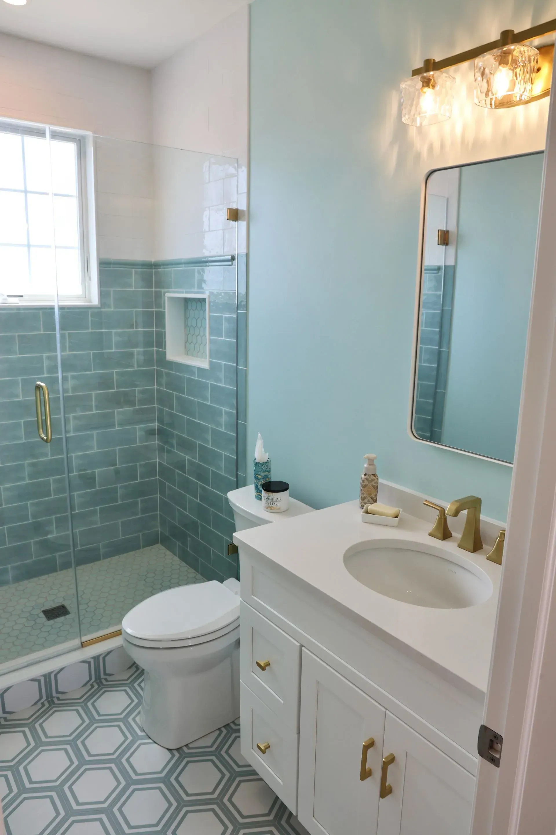 Modern bathroom with light blue walls, a white vanity, a glass-enclosed shower with blue tiles, and hexagon floor tiles.
