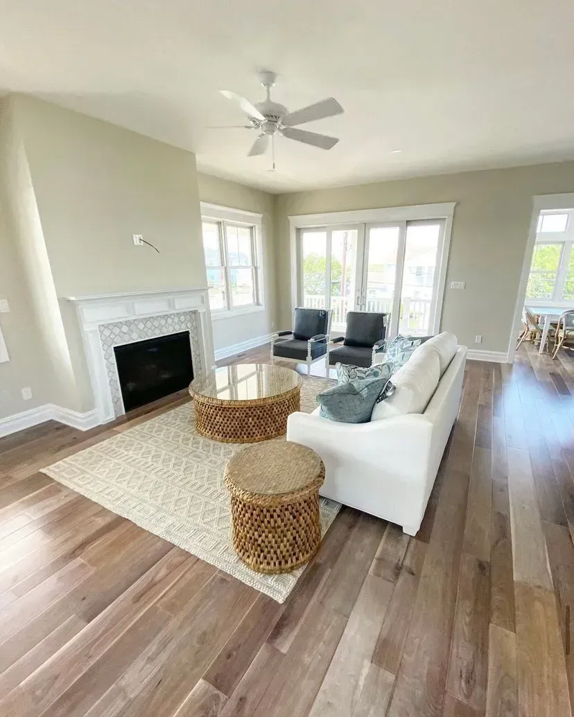 A bright living room with a fireplace, white sofa, two dark armchairs, woven side tables, and wood floors.