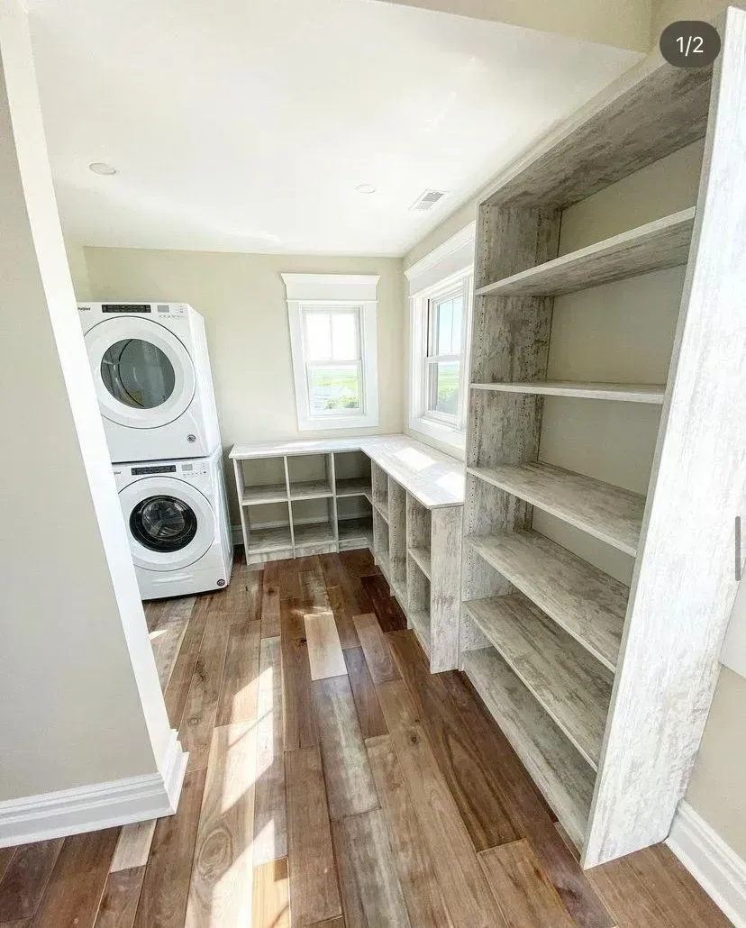 A laundry room with hardwood floors, a stacked washer and dryer, open white cubby shelving, and tall built-in bookshelves.