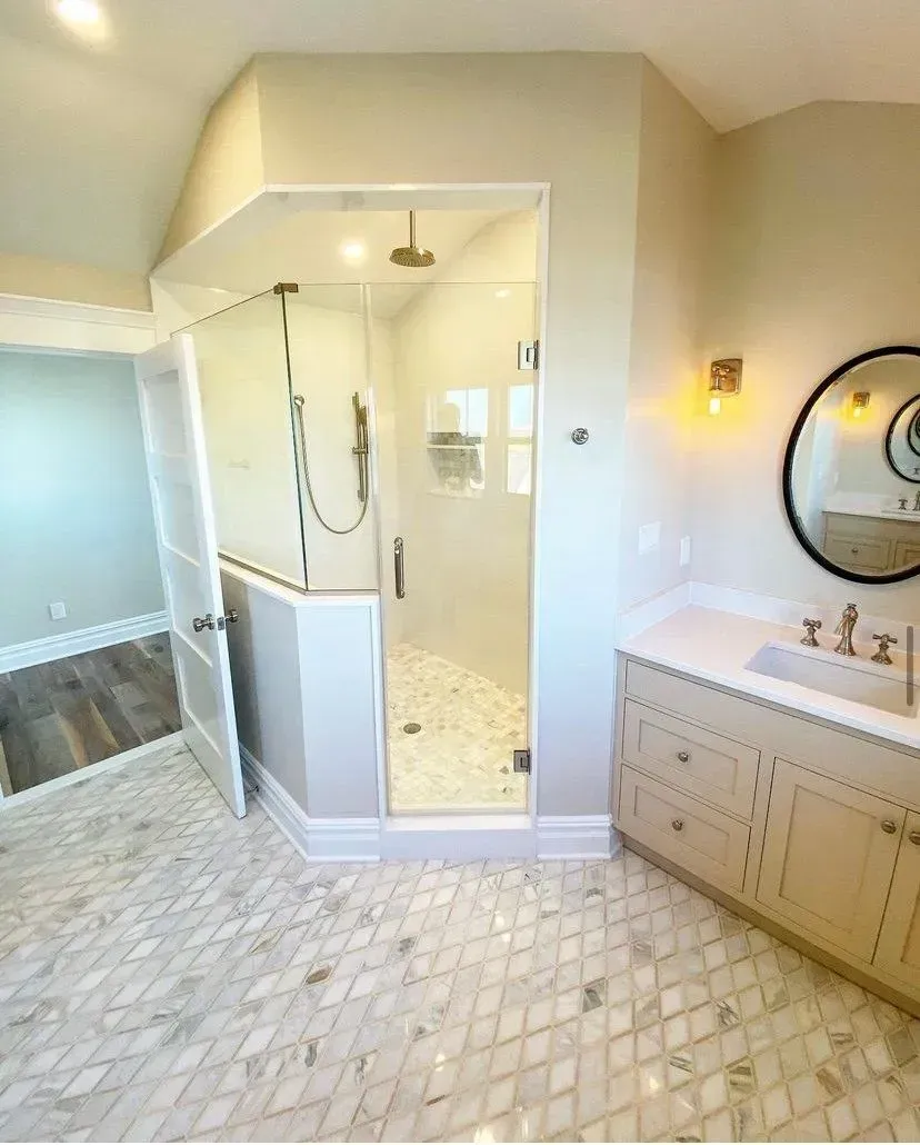 A modern bathroom with a glass-enclosed shower, light-colored vanity, marble flooring, and a round wall mirror.