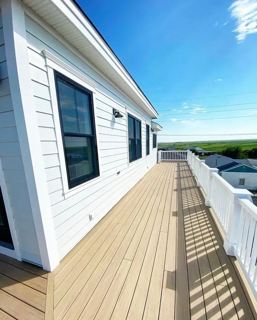 A wide, wooden deck with white railings runs alongside a white house with black-framed windows under a sunny blue sky.
