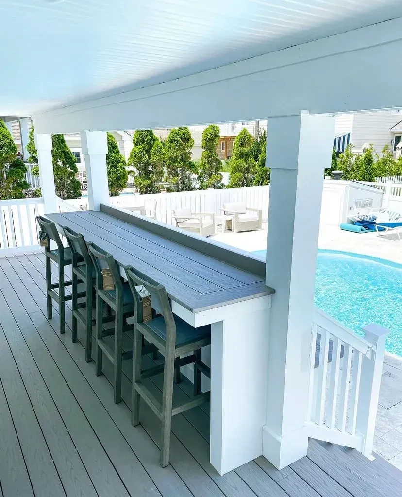 An outdoor bar with stools on a deck overlooking a swimming pool and a white fence.