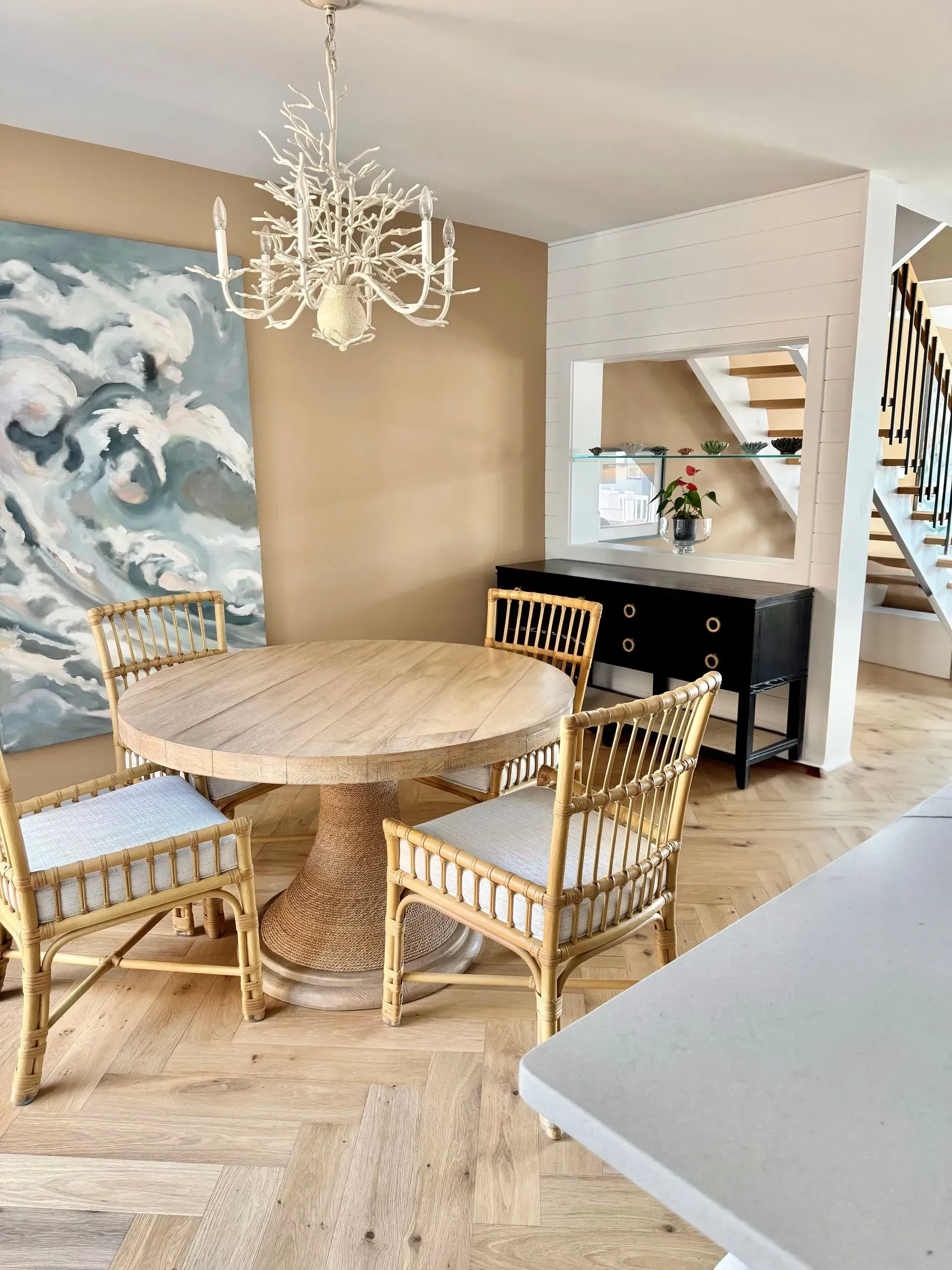 Dining area with a round light wood table, rattan chairs, a chandelier, and a large abstract painting on a tan wall.