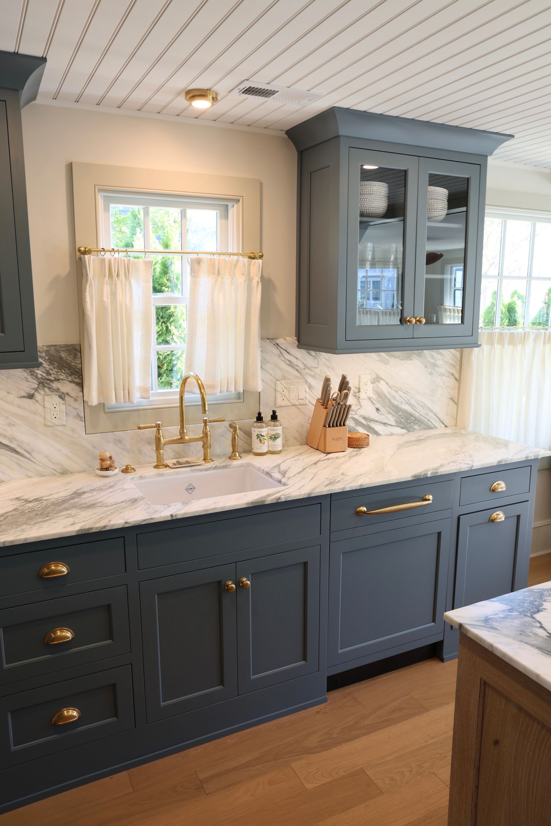 Modern kitchen with navy cabinets, marble countertop, and a glass-front wall cabinet by a sink under a window.