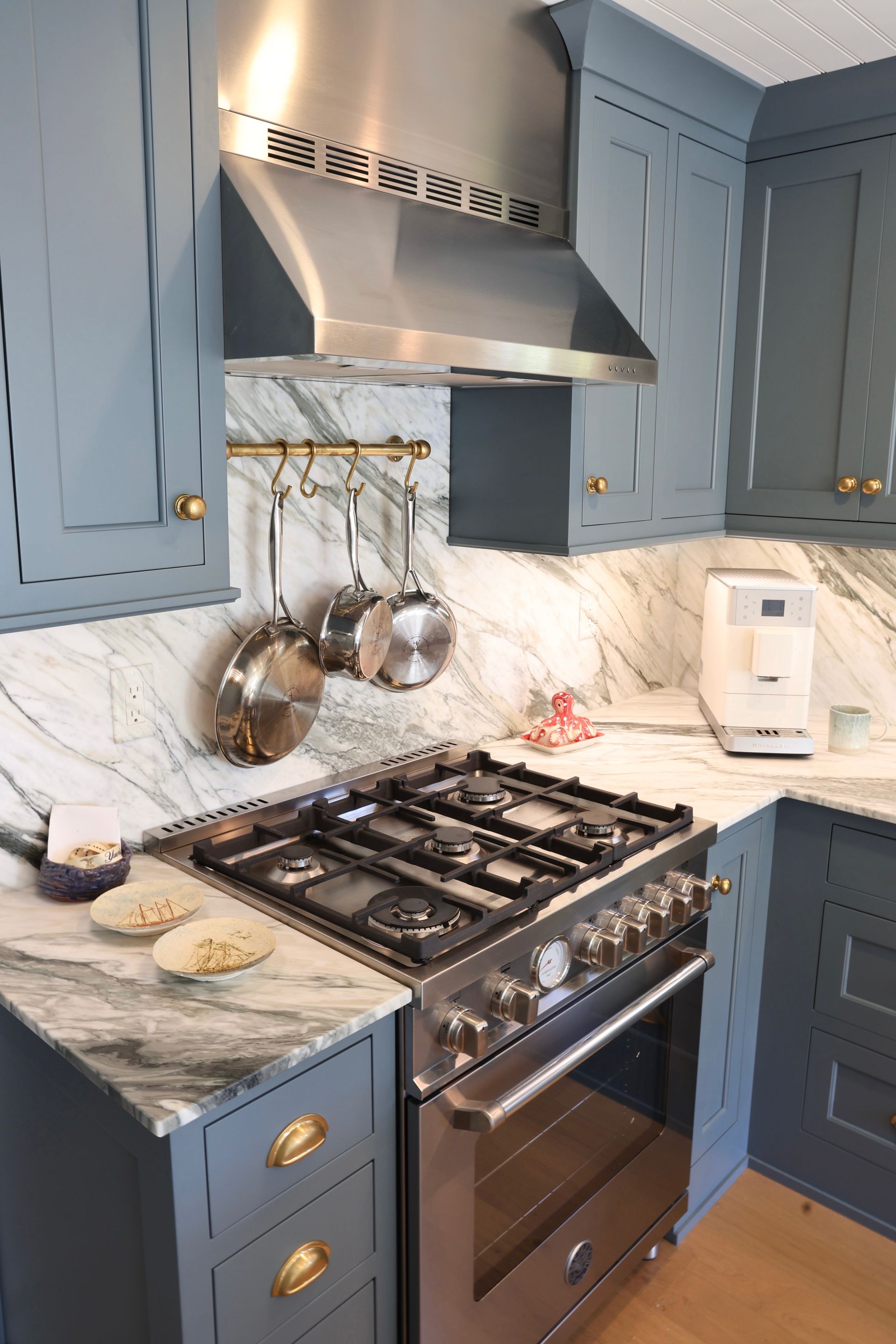 Blue kitchen with marble backsplash, stainless gas stove, hanging pots, and white coffee maker on the counter.
