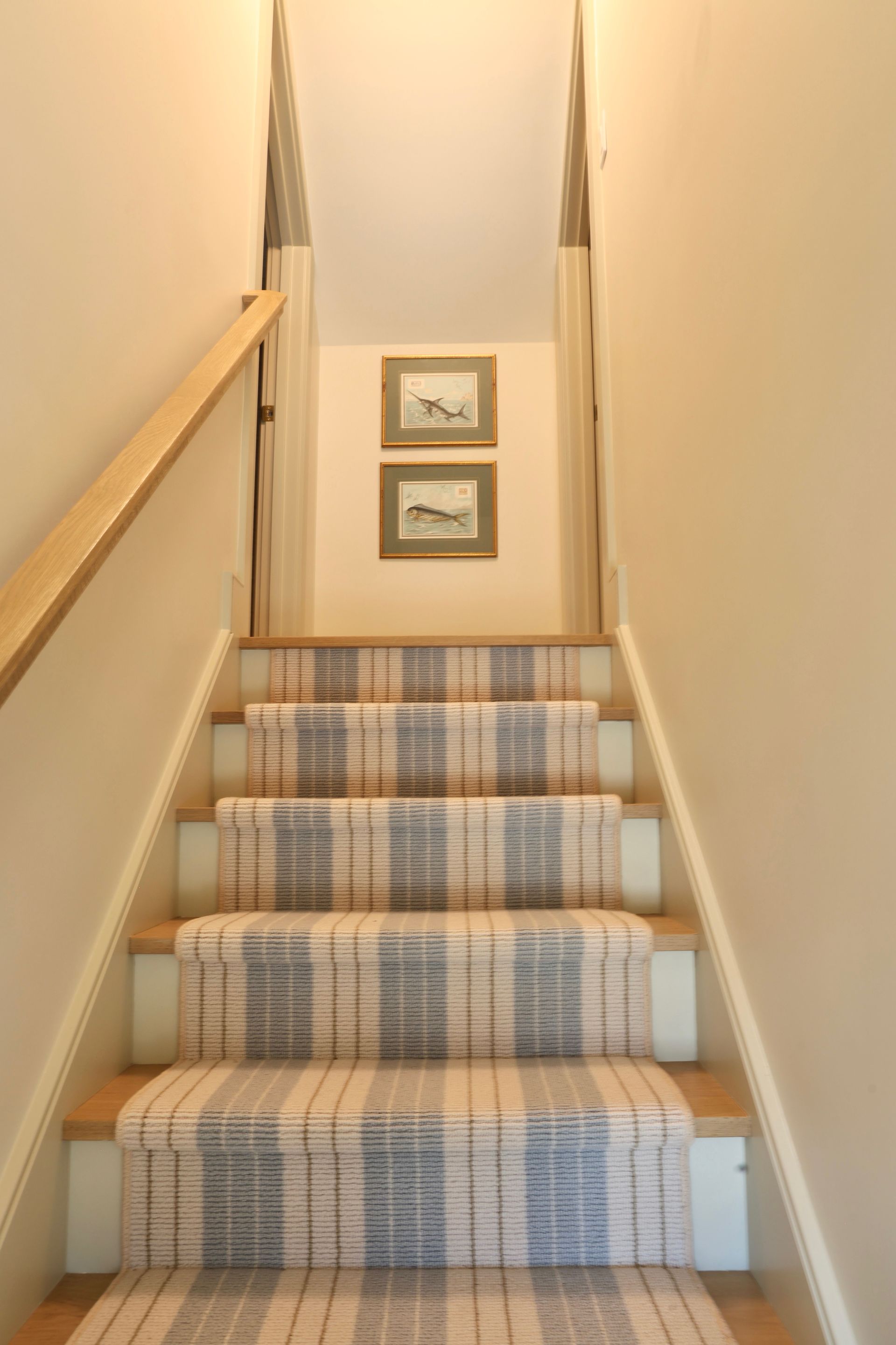 Narrow carpeted staircase with plaid runner, beige walls, and two framed pictures on the landing wall.