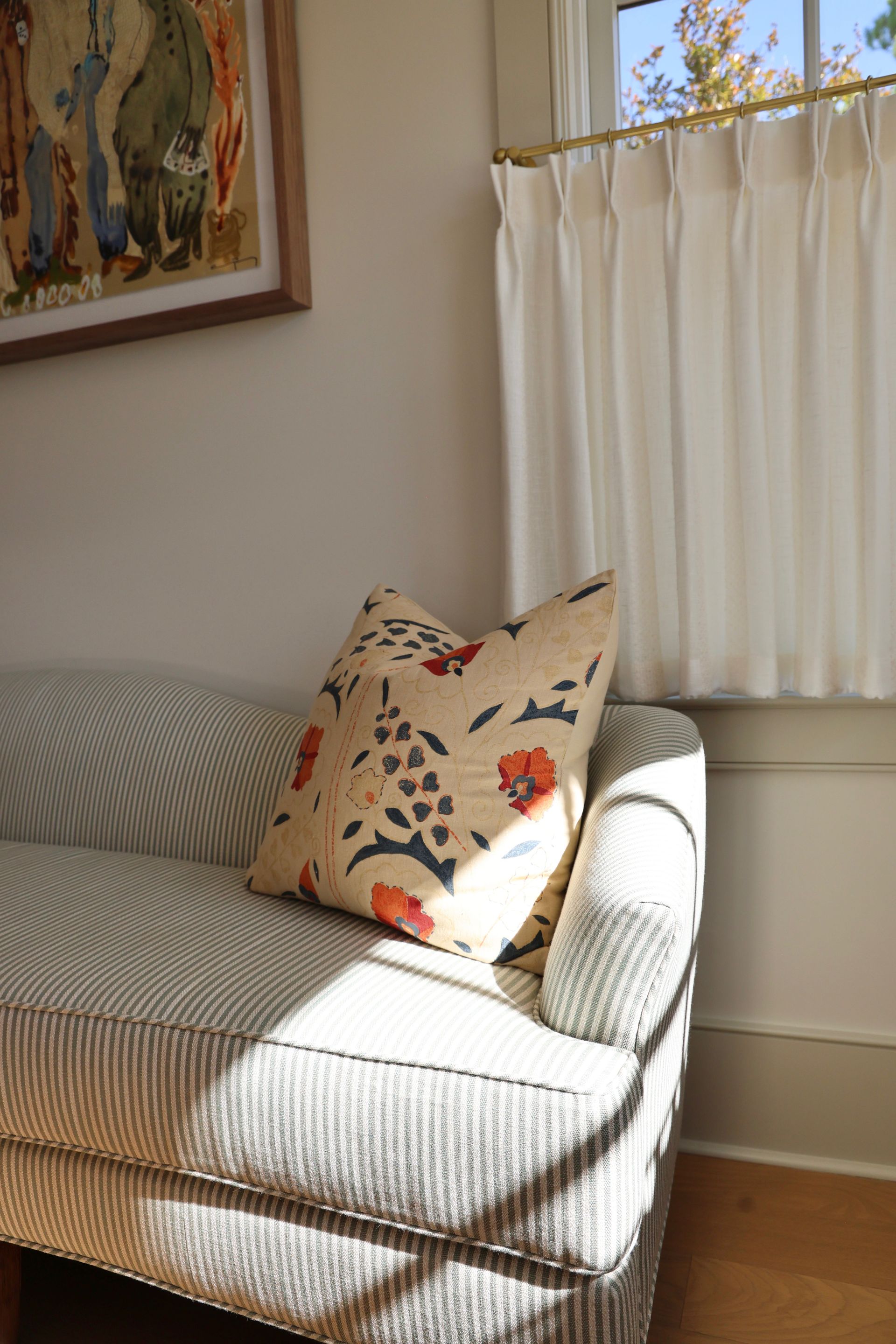 Sunlit sofa corner with patterned cushions and a curtained window beside framed wall art.