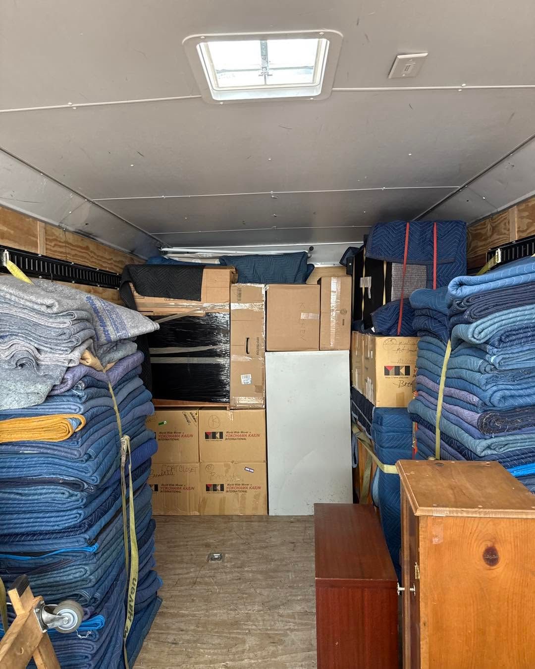 Packed storage area with stacked blue tarps, cardboard boxes, and wooden furniture.