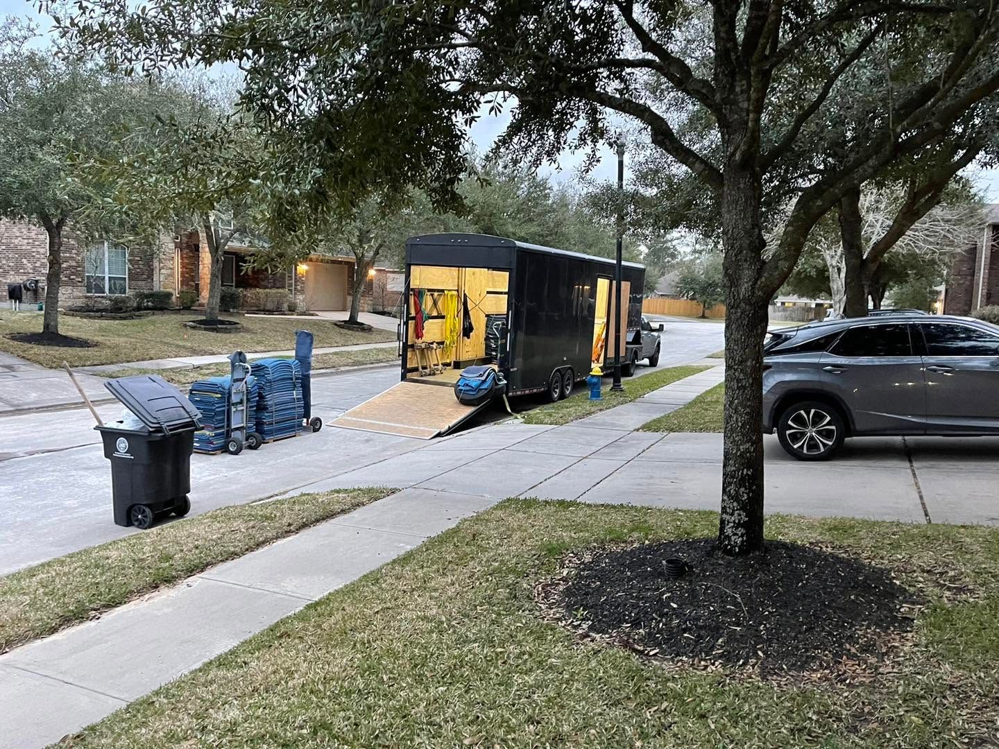 Yellow utility trailer on a suburban sidewalk beside stacked blue water jugs and parked cars under trees