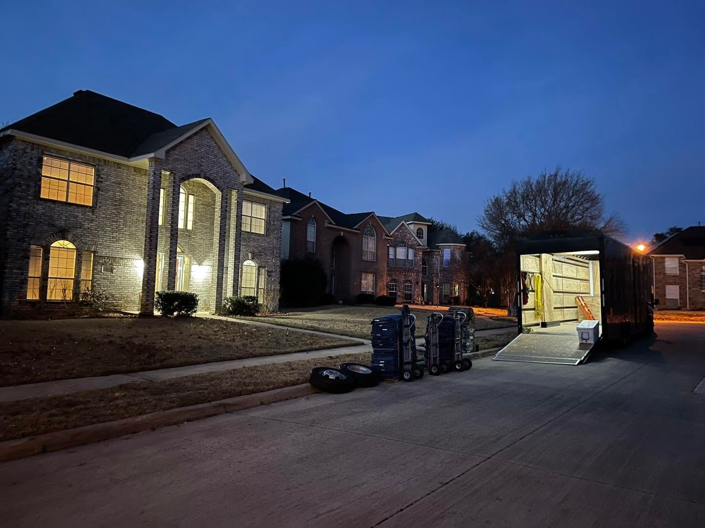 Nighttime exterior of a stone house with lights on, moving truck open, and stacks of boxes on the driveway