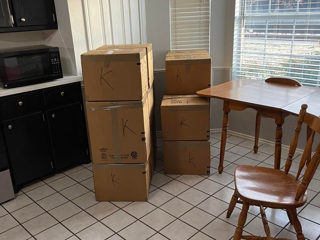 Cardboard boxes stacked in a kitchen next to a table, chair, and black appliances on a tiled floor.