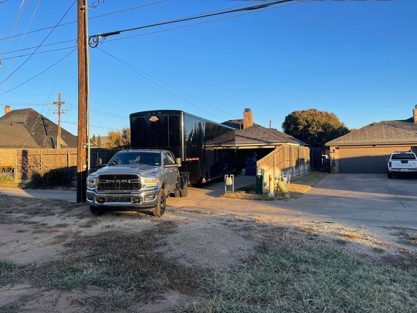 Silver pickup truck in front of a black trailer on a gravel driveway beside brick houses at sunset