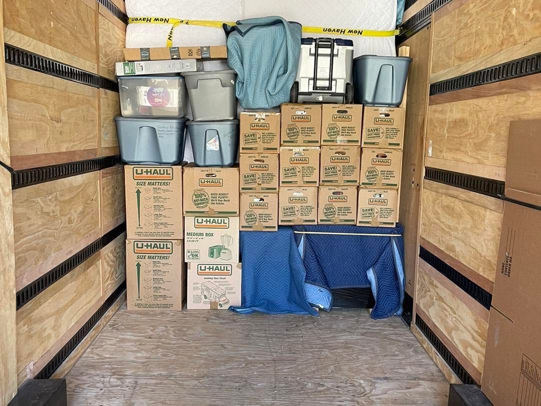 Box-filled moving truck with stacked storage bins, cardboard boxes, and a blue tarp at the back