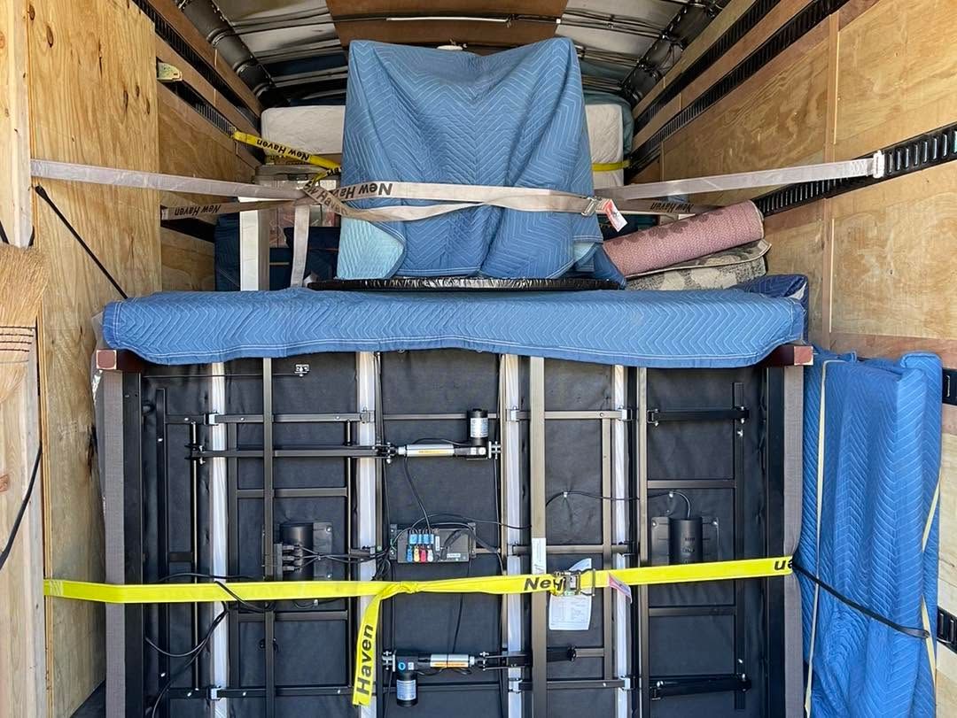 Stacked furniture strapped inside a moving truck with blue padding and yellow tie-down straps.