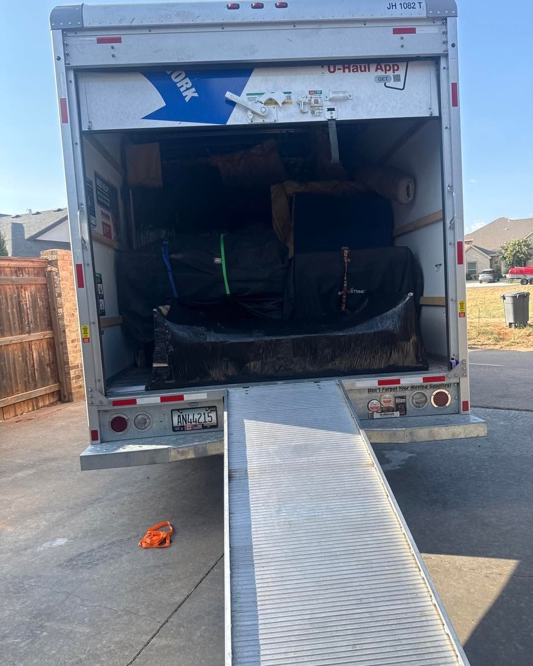 Open truck trailer with folded ramp, carrying dark furniture or equipment inside.