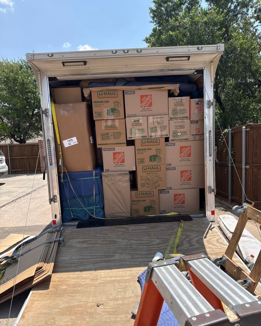 Overloaded moving truck trailer packed with cardboard boxes and a blue bin, rear doors open on a sunny driveway