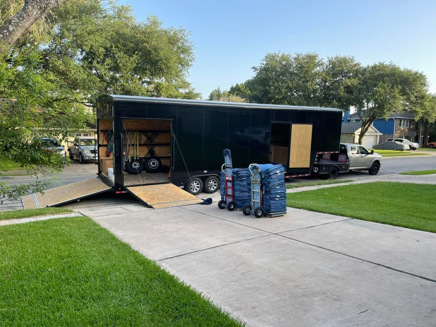 Open trailer unloading blue bottled water cases onto a driveway, with a ramp and trees nearby